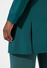 Tunika turchese con spacchi laterali, realizzata in tessuto morbido. Presenta un polsino con bottoni e una texture liscia, abbinata a pantaloni coordinati.