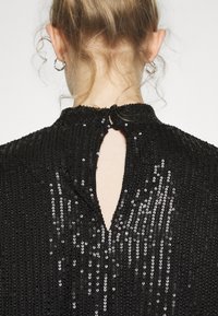 Vue arrière d'une personne aux cheveux blonds attachés en chignon, portant un haut noir à sequins avec une ouverture en trou de serrure dans le dos et une fermeture boutonnée au niveau du cou.