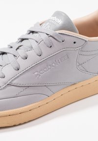 Graue Ledersneaker mit einer strukturierten beigen Gummisohle, ausgestattet mit grauen Schnürsenkeln und einem Logo auf der Zunge. Schlichtes, sauberes Design mit Nähdetails.