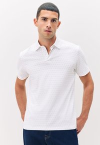 Hombre con cabello corto vestido con una camiseta polo de manga corta blanca con un pequeño patrón, de pie con las manos detrás de la espalda contra un fondo claro y liso.