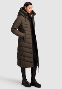 Sieviete, kas ģērbusies garā, olīvu zaļā pildītā virsjakā ar kapuci, melnās legingās un melnās masīvajās zābakos, stāvot pret pelēku fona.