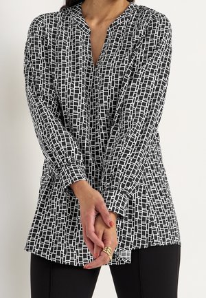 Kvinde iført en langærmet bluse med sort-hvidt geometrisk mønster, guldringe på fingrene, hænderne foldet foran sig, og sorte bukser.