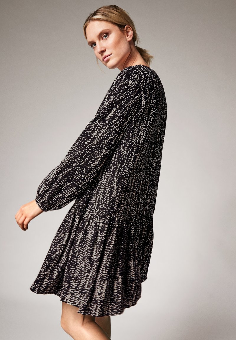 Schwarzes Kleid mit weißem Fleckenmuster, langen Ärmeln, rundem Ausschnitt und gestuftem Rüschensaum. Leichter Stoff mit lockerer Silhouette.