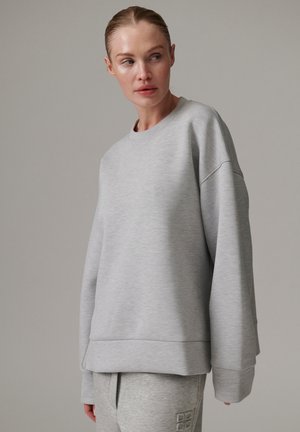 Kvinde iført løs lysegrå sweatshirt og matchende joggingbukser, der kigger sidelæns mod en ensfarvet grå baggrund.
