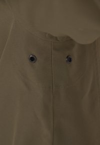 Oliivivihreä kangas, jossa on pyöreitä niittejä kaulan alueella, sileä pinta ja siistit saumat, korostaen minimalistista muotoilua.