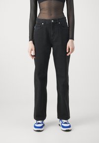 Jeans de mezclilla negra con diseño de pierna recta, con dos bolsillos frontales y cierre de botones. Combinados con un top transparente de manga larga y zapatillas con detalles en azul.