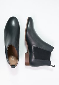 Stivali Chelsea in pelle nera con pannelli elastici laterali e suole in legno marrone, un stivale in posizione verticale e l'altro sdraiato su un lato.