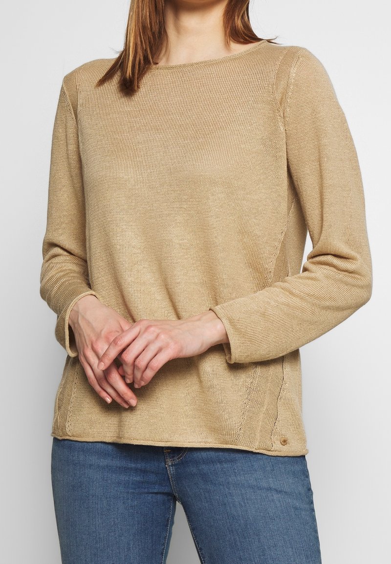 Beige langslijvende gebreide top met een gestructureerde afwerking, ronde halslijn, zijsplitten en een relaxte pasvorm, gedragen over blauwe jeans.