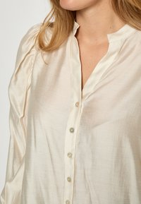 Krēmveida satīna blūze ar stāvošu apkakli, smalku spīdumu, pogu priekšpusi ar iridescentām pogām un pūkām piedurknēm.