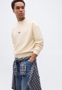 Jeune homme portant un pull crème avec un logo bleu, un jean bleu et une chemise à carreaux bleu marine nouée, regardant vers sa gauche les mains dans les poches.