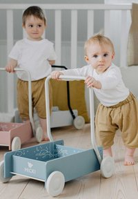 Juguete de empuje de madera en azul con ruedas blancas, con ilustraciones divertidas. Dos niños pequeños vestidos con camisas blancas y pantalones cortos beige.