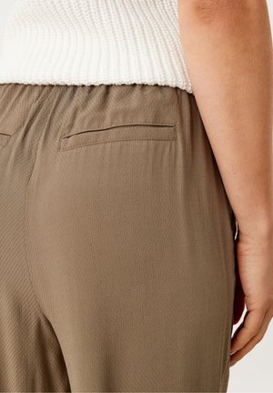 La main droite repose le long du corps, portant un pantalon beige texturé et un haut blanc en maille visible à la taille.