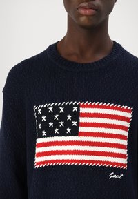 Granatowy sweter z dzianiny z teksturowanym wzorem amerykańskiej flagi na piersi, w kolorach czerwonym, białym i niebieskim, z białymi akcentami haftu.
