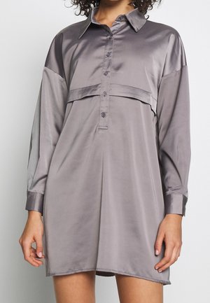 Mujer con un vestido-chemise de satén gris, de manga larga y holgado, con botones y cuello, posando frente a un fondo liso.