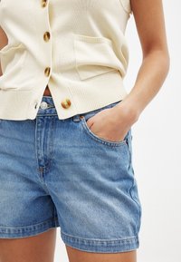 Gai beige adīts kardigans ar zelta krāsas pogām un sānu kabatām, apvienots ar ziliem džinsa šortiem ar tradicionālu piegriezumu un izšūtu apakšmalu.