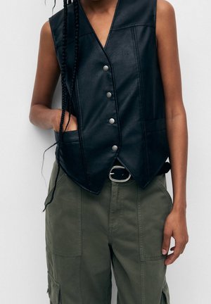 Personne portant un gilet en cuir noir sans manches avec des boutons argentés et un pantalon cargo vert olive avec une boucle de ceinture noire, mains détendues.