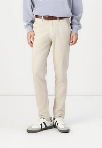 Pantalons en coton beige avec une coupe droite, accompagnés d'une ceinture marron. Portés avec des baskets blanches ornées de rayures noires.