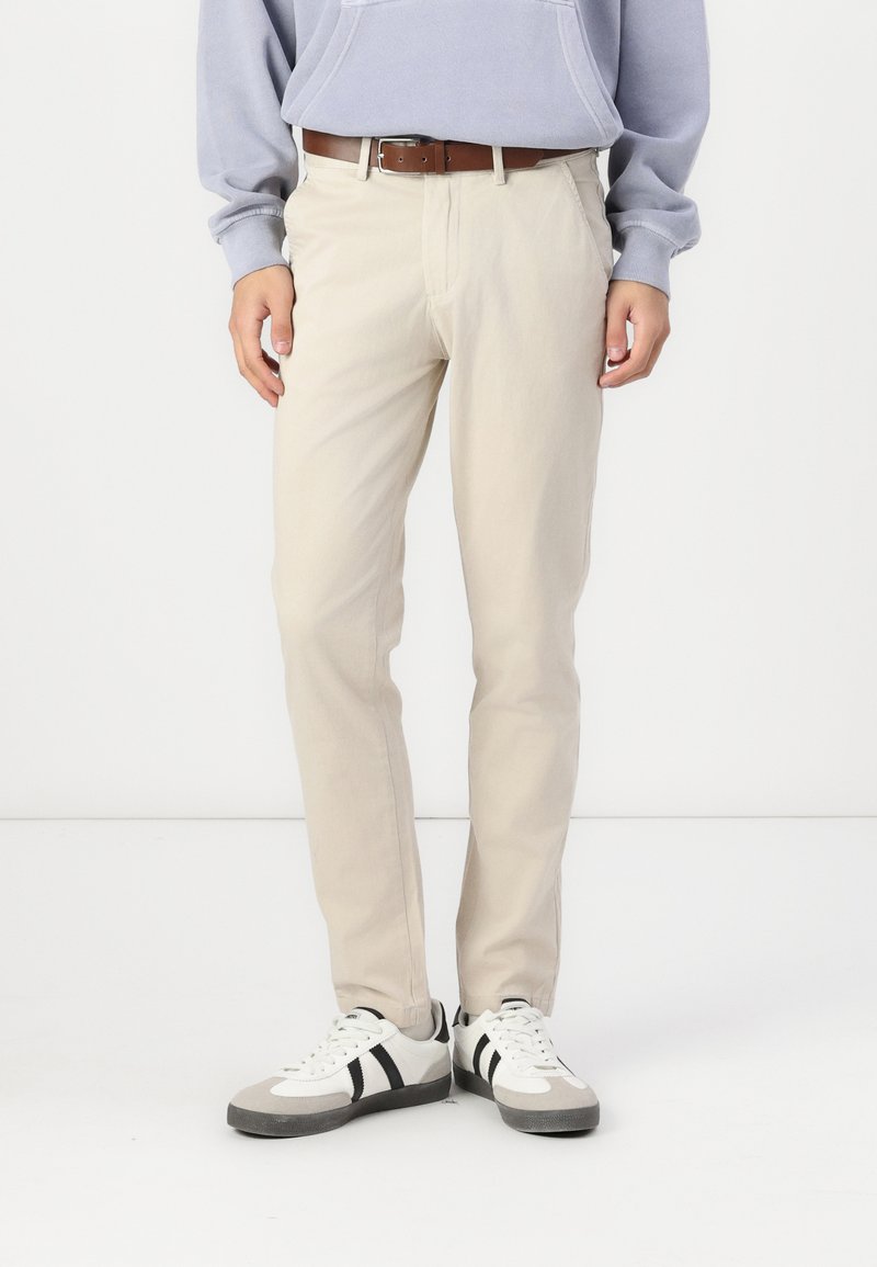 Pantalons en coton beige avec une coupe droite, accompagnés d'une ceinture marron. Portés avec des baskets blanches ornées de rayures noires.