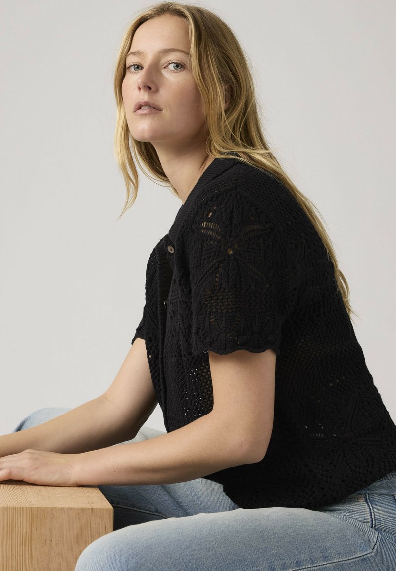 Vrouw met lang blond haar, gekleed in een zwart gehaakt shirt met korte mouwen en lichtblauwe spijkerbroek, zittend met handen rustend op een houten oppervlak.