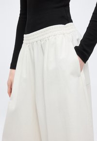 Άτομο που φοράει άνετο λευκό παντελόνι με ελαστική μέση και πλαϊνές τσέπες, συνδυασμένο με στενό μαύρο μακρυμάνικο μπλουζάκι.