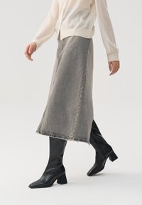 Grijze denim midi-jurk met een rauwe zoom, gecombineerd met zwarte leren blokhaklaarzen, gedragen met een doorschijnend lichtgekleurde top.