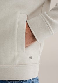 Sudadera de forro polar color crema con un bolsillo lateral y un pequeño logo en el dobladillo. Presenta una tela texturizada y un ajuste relajado.