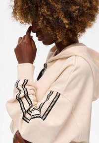 Persoon met krullend haar die een beige hoodie draagt met zwart-wit geborduurde strepen op de mouwen, terwijl ze hun gezicht vlakbij de neus aanraakt.