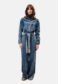 Denim overall met een kraag, lange mouwen, voorzakken en een getailleerde taille. De stof is lichtblauw met een vervaagde textuur.