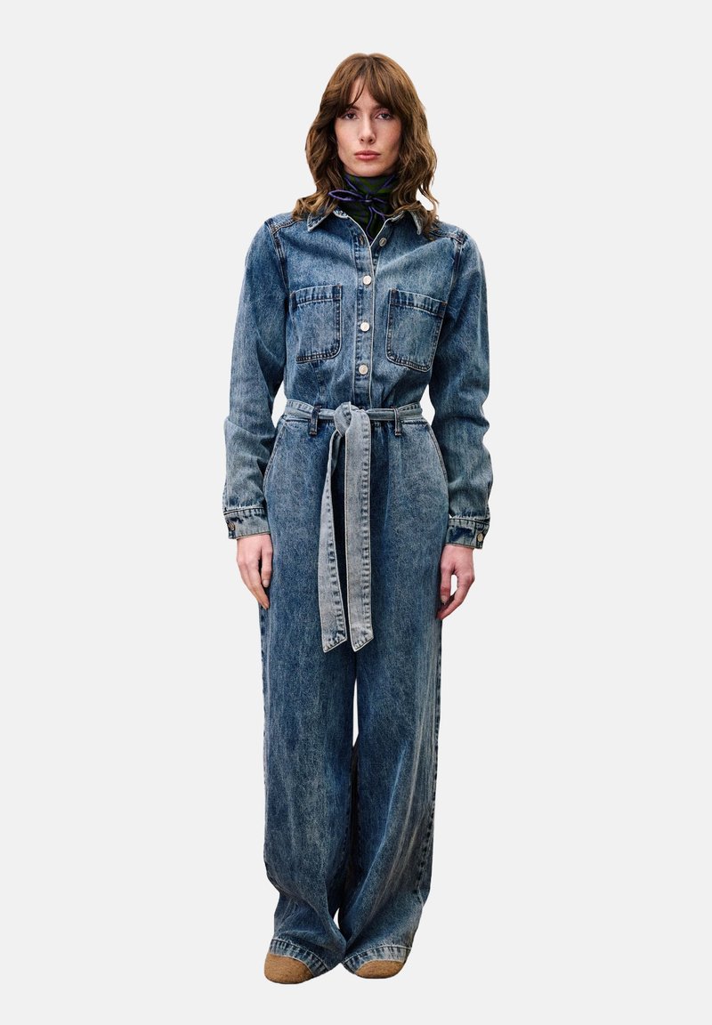 Denim overall met een kraag, lange mouwen, voorzakken en een getailleerde taille. De stof is lichtblauw met een vervaagde textuur.
