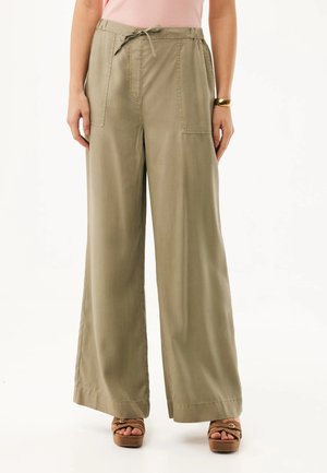 Mujer con pantalones beige de pierna ancha y bolsillos frontales, cintura con cordón, camiseta rosa, pulsera dorada y sandalias de plataforma marrones.