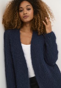 Jūras zilas krāsas pūkains kardigans ar dziļu V veida izgriezumu un atvērtu priekšdaļu, ar garām piedurknēm un mīkstu, teksturētu adījuma materiālu.
