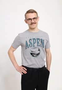 Camiseta de algodón gris con un estampado gráfico azul de "Aspen" y montañas escénicas. Combinada con pantalones negros y un cinturón negro.