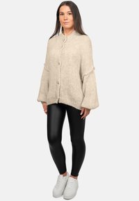 Cardigan beige a maglia con colletto alto e grandi bottoni, abbinato a leggings neri in pelle sintetica e sneakers bianche. Tessuto morbido e testurizzato.