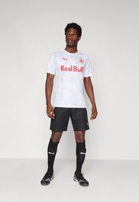 Camisola atlética branca com textura leve, logótipo vermelho "Red Bull", combinada com calções pretos e meias pretas até ao joelho, com a marca Puma.
