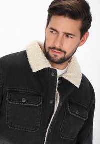 Czarna dżinsowa kurtka z beżowym kołnierzem z sztucznego futra. Zawiera zapinane na guziki kieszenie na piersi oraz metalowe elementy. Materiał wydaje się mocny i teksturowany.