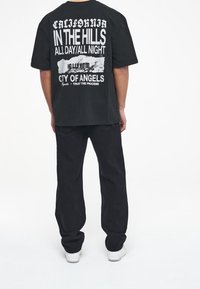 Černé oversized tričko s bílým grafickým textem na zádech, obsahující fráze "Kalifornie" a "Město Andělů". Kombinováno s černými džíny.