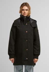 Manteau noir long avec un col haut, fermeture éclair, boutons-pression, poignets côtelés et deux poches frontales. Extérieur lisse avec une coupe structurée.