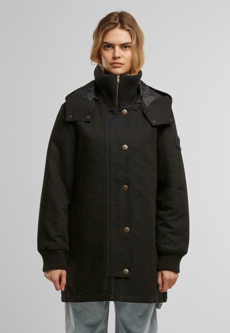Manteau noir long avec un col haut, fermeture éclair, boutons-pression, poignets côtelés et deux poches frontales. Extérieur lisse avec une coupe structurée.