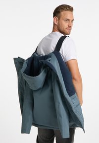 Blå och marinblå isolerad jacka med huva, med tryckknappar och remmar för att bäras på ryggen. Mjuk innervärme synlig.