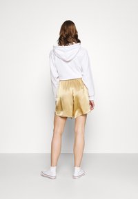 Weißer Hoodie mit Kapuze, kombiniert mit glänzenden goldenen Satin-Shorts, die eine lockere Passform und einen elastischen Bund aufweisen. Weiße High-Top-Schuhe komplettieren das Outfit.