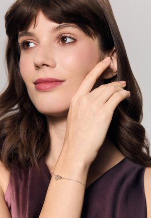 Srebrna bransoletka łańcuchowa z zawieszką w kształcie serca ozdobioną małymi przezroczystymi kamieniami, noszona na nadgarstku, przy którym znajduje się dłoń dotykająca twarzy.