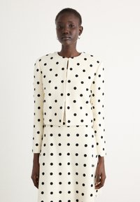 White, textured fabric jacket with black polka dots, cropped design, round neckline, and front closure. Complemented by a matching skirt.