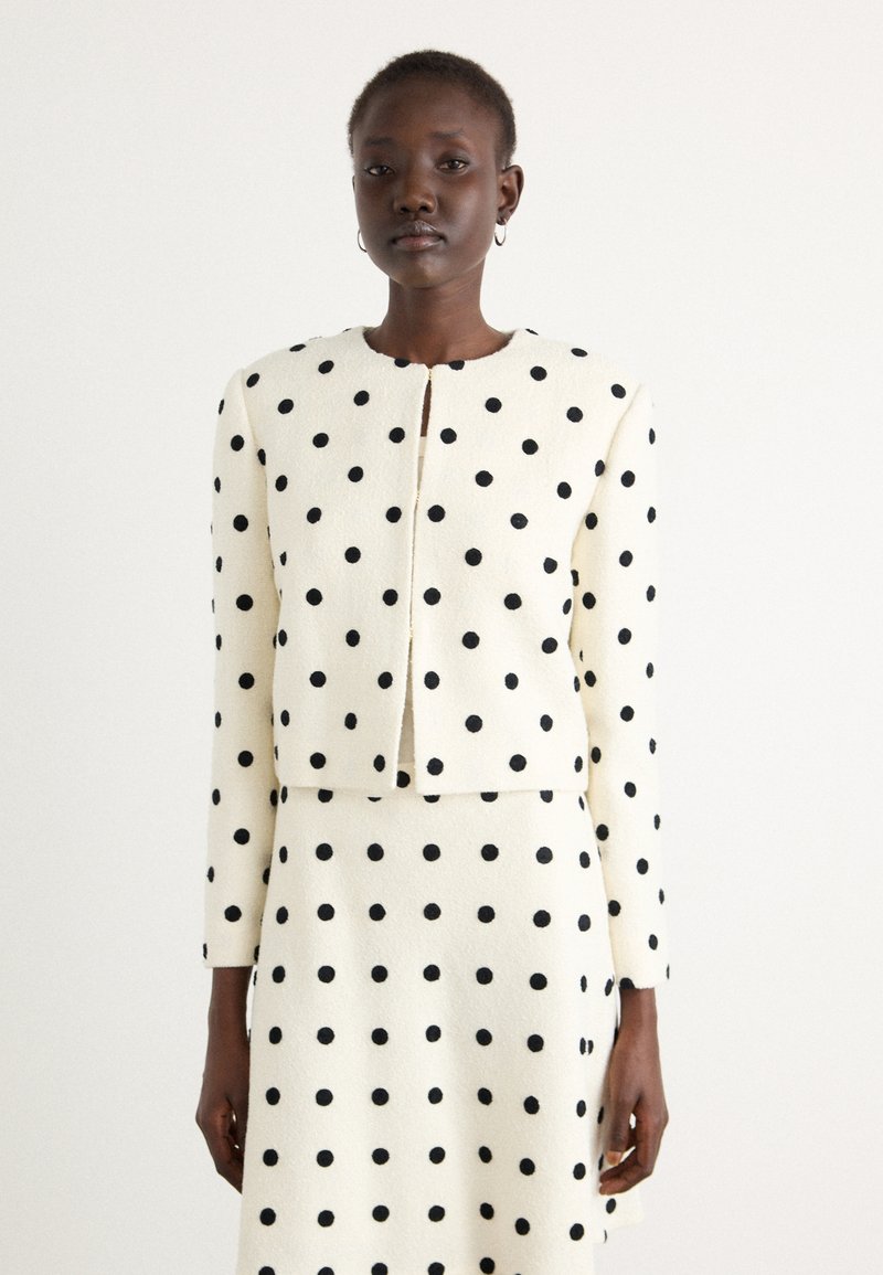 White, textured fabric jacket with black polka dots, cropped design, round neckline, and front closure. Complemented by a matching skirt.