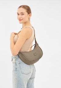 Borsa hobo in tessuto verde oliva, caratterizzata da una forma curvata, zip frontale e patch del logo. Il modello la indossa sopra una maglia beige e jeans azzurri.