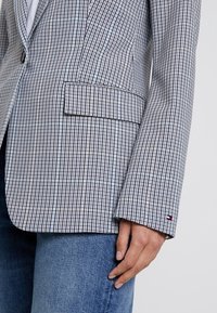 Blazer a scacchi in blu e bianco, caratterizzato da un risvolto, chiusura con un solo bottone e una tasca laterale. Tessuto testurizzato con cuciture visibili.