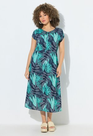 Mujer con cabello rizado vestida con un vestido midi de estampado tropical azul y verde y sandalias beige de plataforma, de pie frente a una pared blanca.