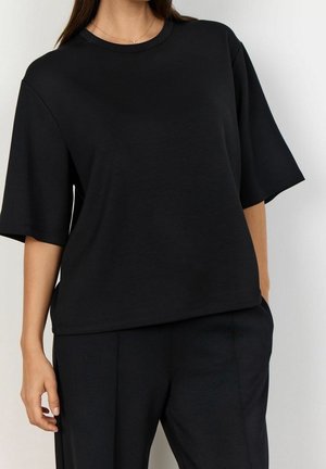 Femme portant un haut noir ample à manches courtes et un pantalon noir assorti, debout avec une main partiellement dans la poche, sur fond blanc.