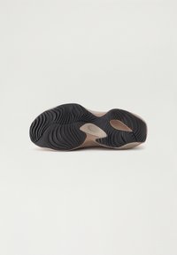 Semelle extérieure d'une basket beige avec une semelle en caoutchouc noire texturée en vagues, montrant des rainures courbes et des éléments de design découpés sur fond blanc.