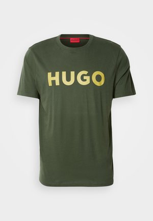T-shirt en coton vert olive avec un col rond et des manches courtes. Présente un grand texte doré et en gras "HUGO" sur le devant.
