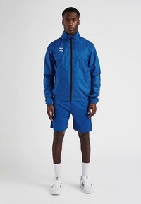 Blå sportjacka med dragkedja och elastiska muddar; matchas med blå shorts. Modellen står mot en neutral bakgrund, iklädd vita strumpor och skor.